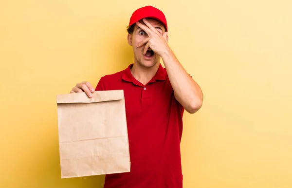 young handsome guy looking shocked, scared or terrified, covering face with hand. delivery and take away concept