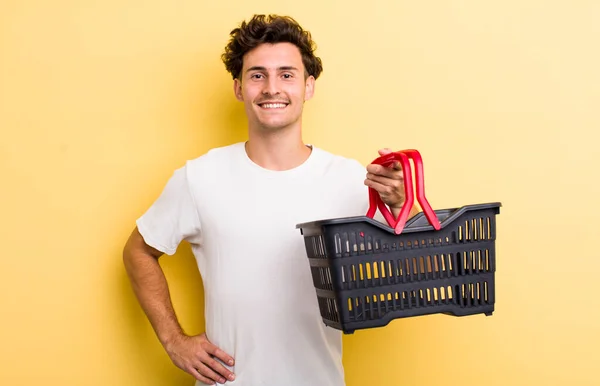 若いハンサムな男が喜んで笑顔で手を腰と自信を持って 空のショッピングバスケットのコンセプト — ストック写真