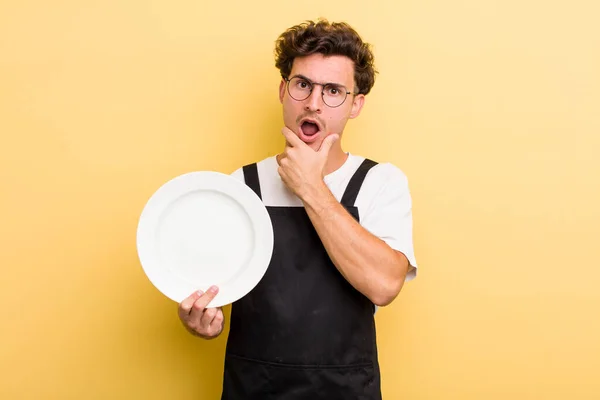 young handsome guy with mouth and eyes wide open and hand on chin. empty dish concept