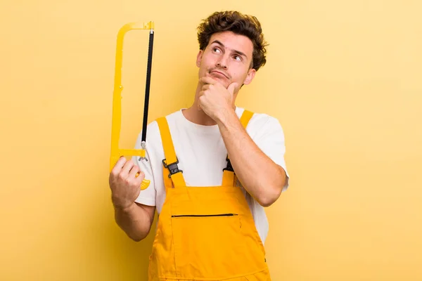 young handsome guy thinking, feeling doubtful and confused. handyman and a saw concept