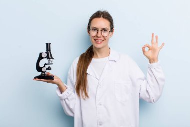 Genç yetişkin bir kadın kendini mutlu hissediyor. Onayını iyi bir jestle gösteriyor. Mikroskoplu bir bilim adamı