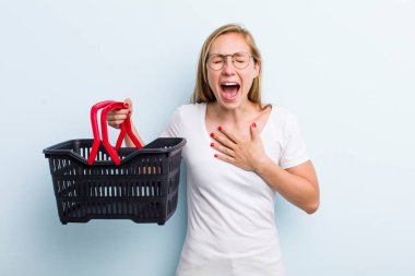 Sarışın, genç, yetişkin bir kadın komik bir şakaya yüksek sesle gülüyor. Alışveriş sepeti