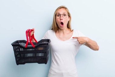 Şok olmuş ve ağzı açık, kendini işaret eden genç sarışın bir kadın. Alışveriş sepeti