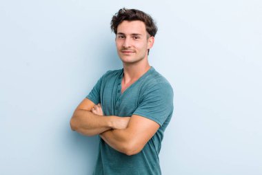 Genç yakışıklı adam kameraya çapraz kollarıyla gülümsüyor ve mutlu, kendinden emin, tatmin olmuş bir ifade, yan bakış.
