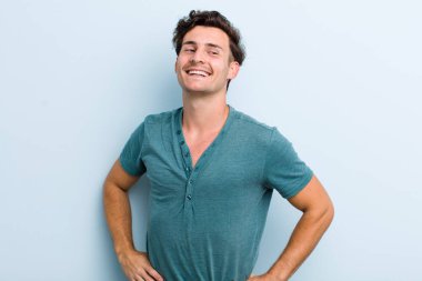 Genç yakışıklı adam mutlu, neşeli ve kendinden emin görünüyor. Gururla gülümsüyor ve iki elini de kalçasında istiyor.