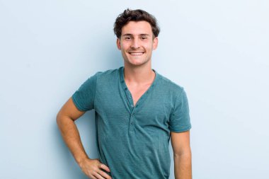 Genç yakışıklı adam mutlu bir şekilde gülümsüyor, eli kalçasında ve kendinden emin, olumlu, gururlu ve arkadaş canlısı.