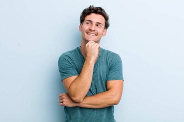 Genç yakışıklı adam mutlu bir şekilde gülümsüyor ve hayal kuruyor ya da şüphe ediyor.