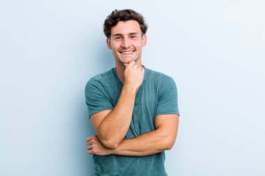 Genç yakışıklı adam mutlu görünüyor ve gülümsüyor. Elleri çenesinde, merak ediyor ya da soru soruyor. Seçenekleri karşılaştırıyor.