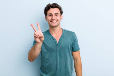 Genç yakışıklı adam gülümsüyor ve mutlu görünüyor, kaygısız ve olumlu, zafer veya barış işareti yapıyor.