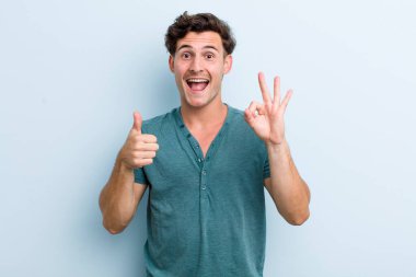 Genç yakışıklı adam kendini mutlu, şaşkın, tatmin olmuş ve şaşırmış hissediyor, tamam gösteriyor ve el kol hareketleri yapıyor, gülümsüyor.