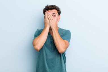 Genç yakışıklı adam elleriyle yüzünü kapatıyor, parmaklarının arasından şaşkın bir ifadeyle yan tarafa bakıyor.