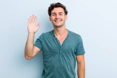 Genç yakışıklı adam mutlu ve neşeyle gülümsüyor, el sallıyor, seni selamlıyor ve selamlıyor, ya da hoşça kal diyor.