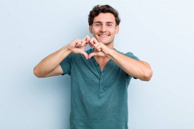 Genç yakışıklı adam gülümsüyor ve mutlu, sevimli, romantik ve aşık, iki eliyle kalbi şekillendiriyor.