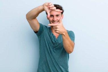 Genç yakışıklı adam mutlu, arkadaş canlısı ve pozitif hissediyor, gülümsüyor ve elleriyle bir portre veya fotoğraf çerçevesi yapıyor.