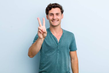 Genç yakışıklı adam gülümsüyor ve arkadaş canlısı görünüyor, iki ya da iki numarayı gösteriyor, elleri önde, geriye doğru sayıyor.