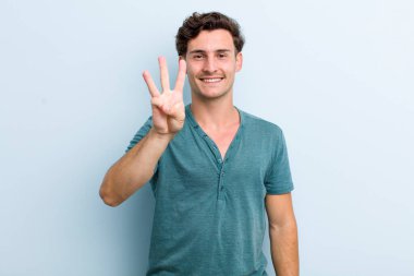 Genç yakışıklı adam gülümsüyor ve arkadaş canlısı görünüyor, üç ya da üç numarayı gösteriyor, eli önde, geriye doğru sayıyor.