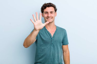 Genç yakışıklı adam gülümsüyor ve arkadaş canlısı görünüyor, 5 ya da 5 numarayı gösteriyor, eli önde, geriye doğru sayıyor.