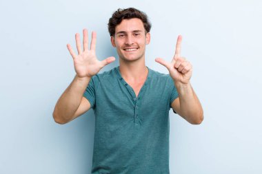 Genç yakışıklı adam gülümsüyor ve dost canlısı görünüyor, 7 ya da 7 numarayı gösteriyor, eli önde, geriye doğru sayıyor.