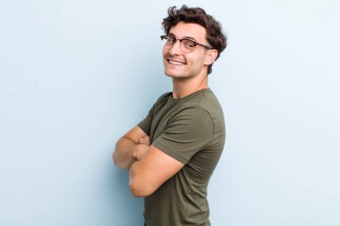 Genç yakışıklı adam neşeyle gülümsüyor, mutlu, tatmin olmuş ve rahatlamış hissediyor, kollarını kavuşturup yan tarafa bakıyor.