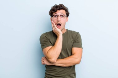 Genç yakışıklı adam şok ve inançsızlık içinde, elleri yanak ve kolları çapraz, sersemlemiş ve şaşırmış hissediyor.