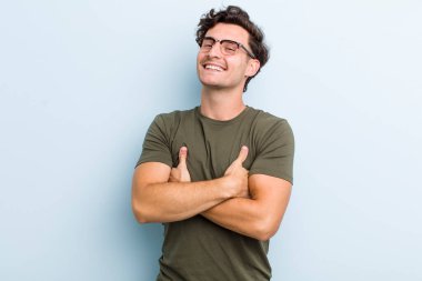 Genç yakışıklı adam kollarını kavuşturup mutlu bir şekilde gülüyor, rahat, pozitif ve tatmin edici bir pozla.