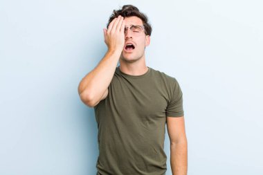 Genç yakışıklı adam uykulu, sıkılmış ve esniyor, baş ağrısı ve yüzünün yarısını kaplayan bir elle.