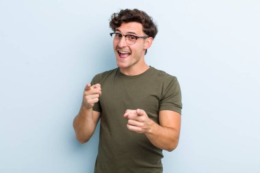 Olumlu, başarılı, mutlu bir tavırla gülümseyen genç yakışıklı adam kamerayı işaret ediyor, elleriyle silah işareti yapıyor.