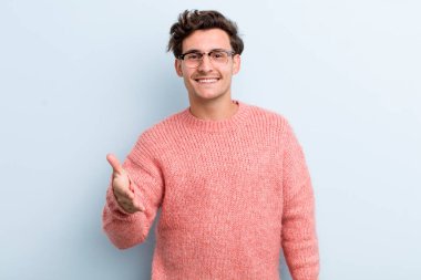 Genç yakışıklı adam gülümsüyor, mutlu görünüyor, kendinden emin ve arkadaş canlısı, bir anlaşmaya varmak için el sıkışıyor, işbirliği yapıyor.