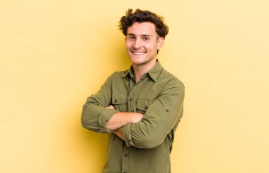 Genç yakışıklı adam kameraya çapraz kollarıyla gülümsüyor ve mutlu, kendinden emin, tatmin olmuş bir ifade, yan bakış.