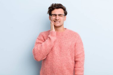 Genç yakışıklı adam yanağını tutuyor ve ağrılı diş ağrısı çekiyor, hasta, sefil ve mutsuz hissediyor, dişçi arıyor.