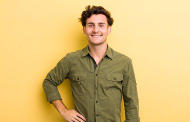 Genç yakışıklı adam mutlu bir şekilde gülümsüyor, eli kalçasında ve kendinden emin, olumlu, gururlu ve arkadaş canlısı.