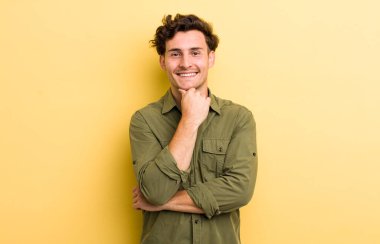 Genç yakışıklı adam mutlu görünüyor ve gülümsüyor. Elleri çenesinde, merak ediyor ya da soru soruyor. Seçenekleri karşılaştırıyor.