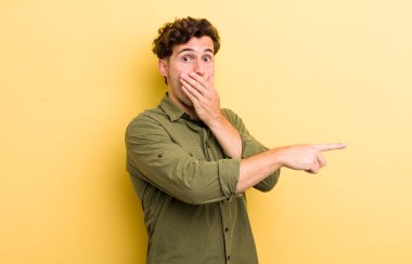 Genç yakışıklı adam kendini mutlu, şaşkın ve şaşırmış hissediyor. Ağzını eliyle kapatıyor ve yanal kopya alanını işaret ediyor.