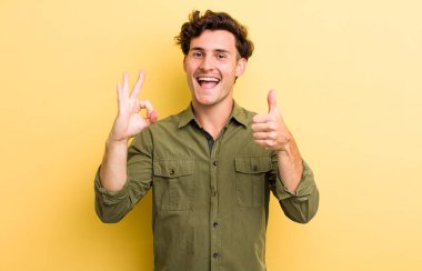 Genç yakışıklı adam kendini mutlu, şaşkın, tatmin olmuş ve şaşırmış hissediyor, tamam gösteriyor ve el kol hareketleri yapıyor, gülümsüyor.