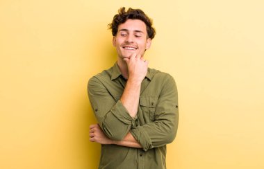 Genç ve yakışıklı bir adam mutlu, kendinden emin bir ifadeyle gülümsüyor. Elleri çenesinde, merak ediyor ve yan tarafa bakıyor.