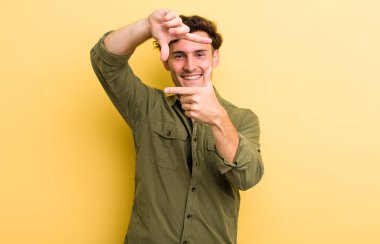 Genç yakışıklı adam mutlu, arkadaş canlısı ve pozitif hissediyor, gülümsüyor ve elleriyle bir portre veya fotoğraf çerçevesi yapıyor.