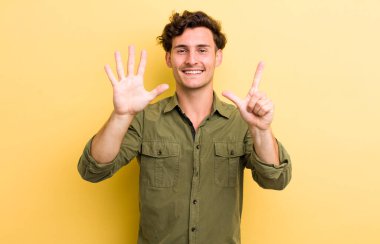 Genç yakışıklı adam gülümsüyor ve dost canlısı görünüyor, 7 ya da 7 numarayı gösteriyor, eli önde, geriye doğru sayıyor.