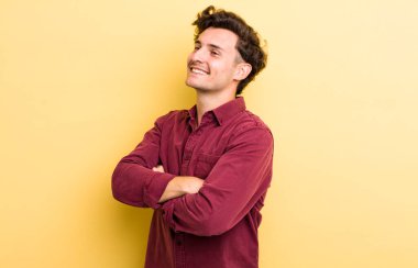 Genç yakışıklı adam neşeyle gülümsüyor, mutlu, tatmin olmuş ve rahatlamış hissediyor, kollarını kavuşturup yan tarafa bakıyor.