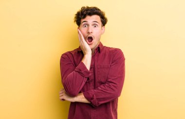 Genç yakışıklı adam şok ve inançsızlık içinde, elleri yanak ve kolları çapraz, sersemlemiş ve şaşırmış hissediyor.