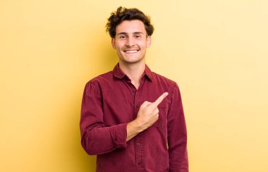 Genç yakışıklı adam neşeyle gülümsüyor, mutlu hissediyor ve kenarı ve yukarıyı işaret ediyor, fotokopi uzayındaki nesneleri gösteriyor.