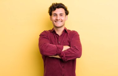 Genç yakışıklı adam mutlu, gururlu ve tatmin olmuş görünüyor. Kollarını kavuşturup gülümsüyor.