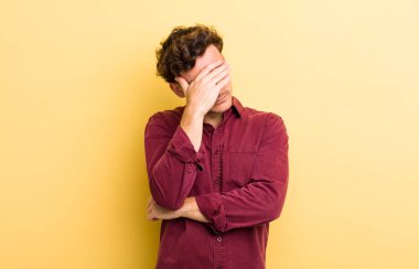 Genç ve yakışıklı bir adam stresli, utanmış ya da üzgün görünüyor, baş ağrısı var, yüzünü eliyle kapatıyor.