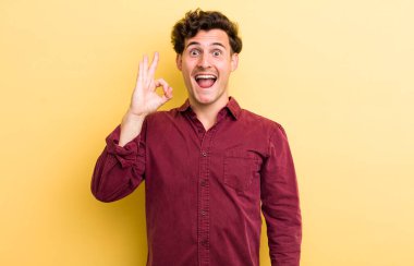 Genç yakışıklı adam kendini başarılı ve tatmin olmuş hissediyor, ağzı açık gülümsüyor, elle işaret veriyor.