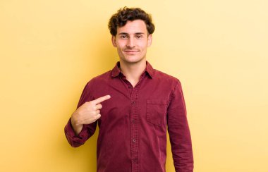 Genç yakışıklı, gururlu, kendinden emin ve mutlu görünüyor, gülümsüyor ve kendini işaret ediyor ya da bir numara işareti yapıyor.