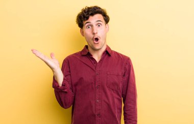 Genç yakışıklı adam şaşırmış ve şoka uğramış görünüyor, çenesi düşmüş, eli açık bir nesneyi tutuyor.