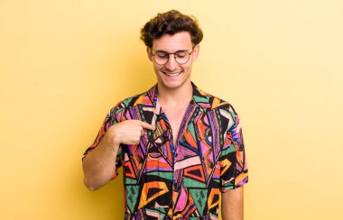 Genç yakışıklı adam neşeyle gülümsüyor, yere bakıyor ve göğsüne işaret ediyor.