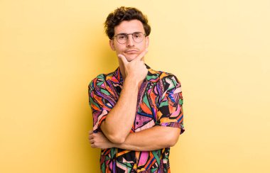 Genç yakışıklı adam ciddi, düşünceli ve güvensiz görünüyor, bir kolu çapraz ve çene üzerinde, seçenekleri tartıyor