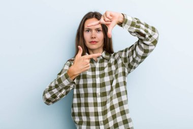 Genç ve güzel bir kadın mutlu, arkadaş canlısı ve pozitif hissediyor, gülümsüyor ve elleriyle bir portre veya fotoğraf çerçevesi yapıyor.