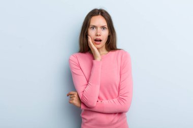young adult pretty woman open-mouthed in shock and disbelief, with hand on cheek and arm crossed, feeling stupefied and amazed