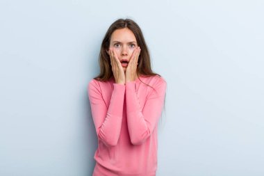 young adult pretty woman feeling shocked and scared, looking terrified with open mouth and hands on cheeks